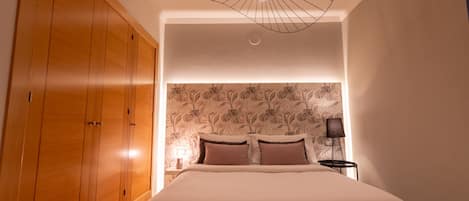 Une chambre avec une grande armoire en bois, un lit doté d’une tête de lit recouverte d’un papier peint à motifs floraux et un luminaire moderne au plafond.