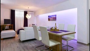 A modern living room with a white sofa, a dining table with four chairs, a TV, and a painting on the wall. - Deluxe Apartments Goya (Zadar)
