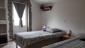 A bedroom with two beds, a stone wall, a window with curtains, and a wooden shelf above the beds.
