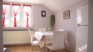 A dining area with a table set for four, white chairs, a window with red curtains, a radiator, and a refrigerator.