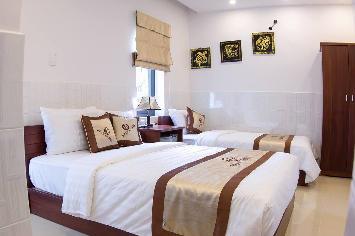 A hotel room with two beds, each with white bedding and brown accents, a bedside table with a lamp, and a window with beige curtains.