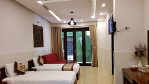 A hotel room with two beds, a TV, a desk with a vase, and a view of greenery outside. - Bean Grass Homestay (Da Nang)