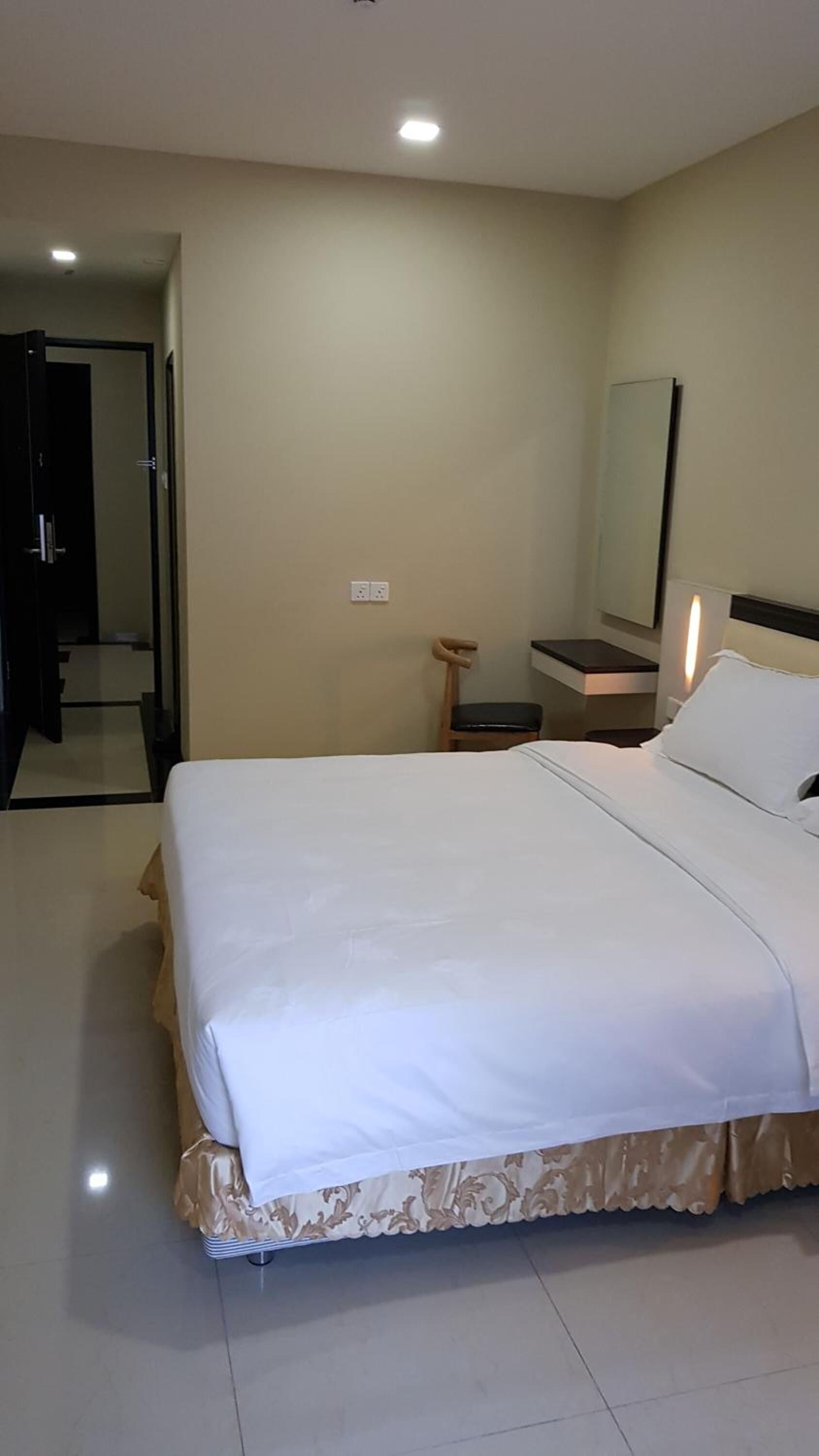 A hotel room with a large bed, a bedside table, a chair, a mirror, and a door leading to another room.