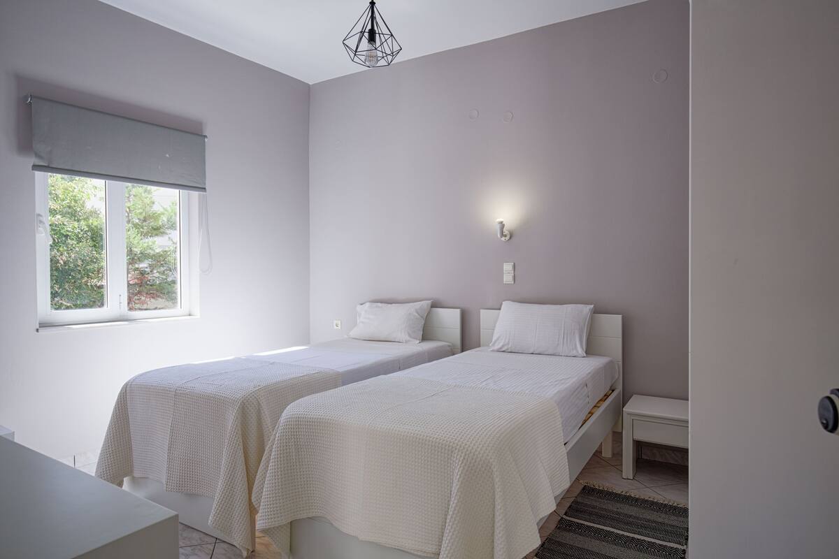 A bedroom with two single beds, a window with a roller blind, a bedside table, and a hanging light fixture.