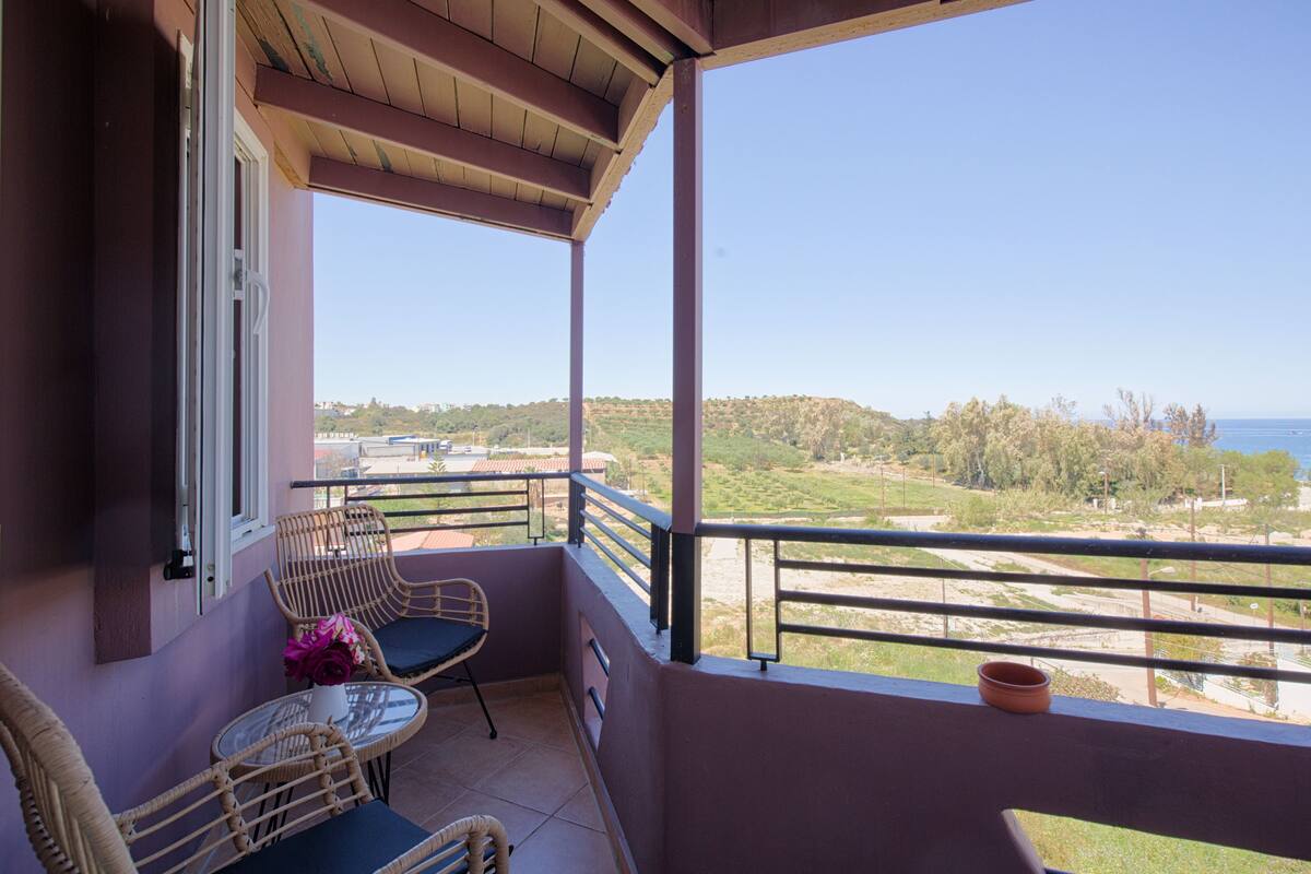 A balcony with wicker chairs and a table, overlooking a scenic landscape with hills and a body of water.