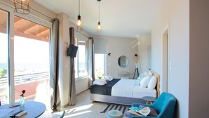 Una habitación de hotel moderna con balcón, una cama con ropa blanca, un sillón azul y una pequeña mesa redonda.