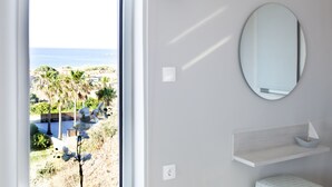 Habitación con un amplio ventanal que ofrece vistas al mar y palmeras. Hay un espejo redondo en la pared y un pequeño estante con un objeto decorativo.