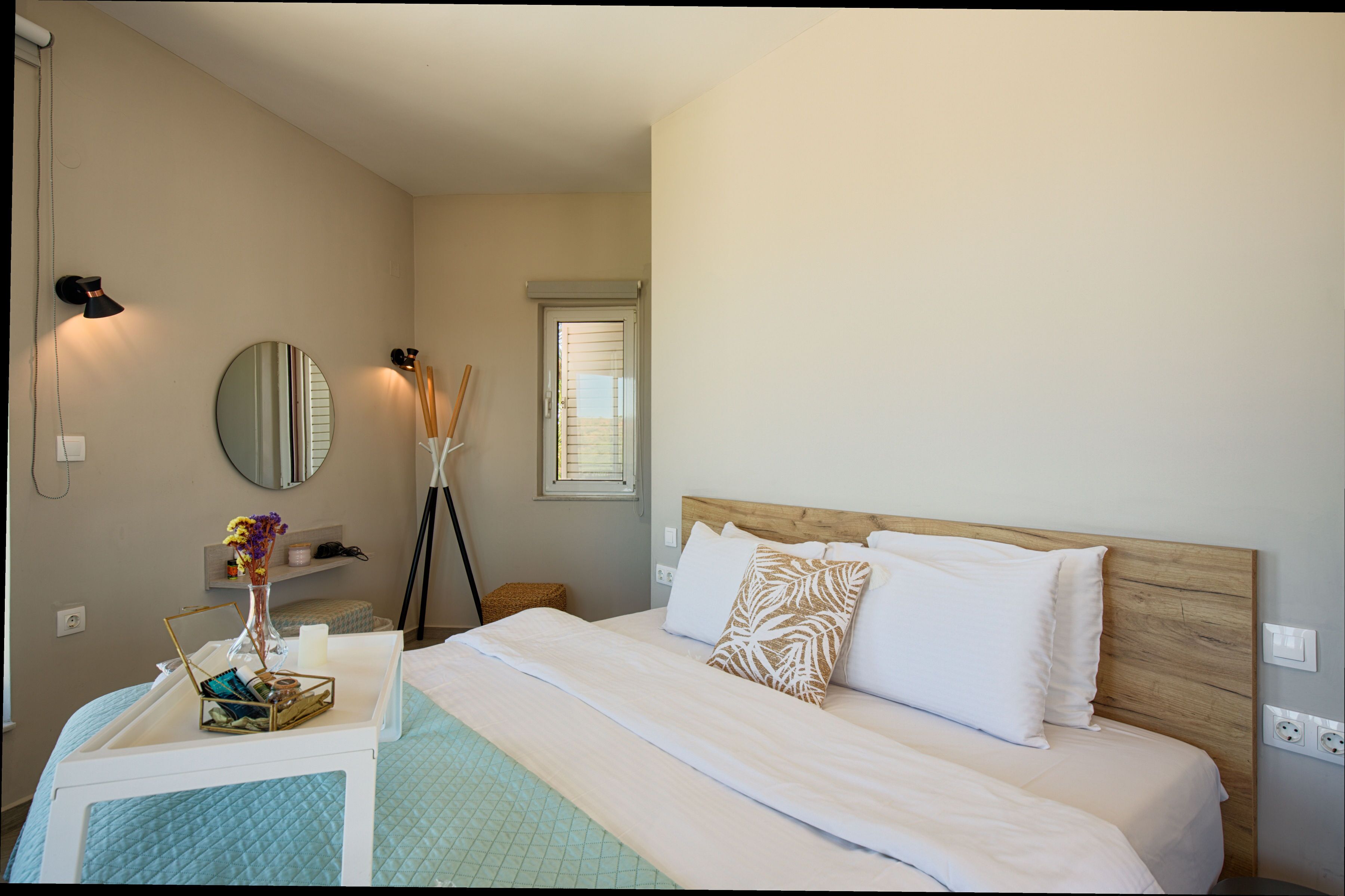 Una habitación de hotel con cama, mesita de noche, lámpara, espejo, ventana y un pequeño taburete de madera.