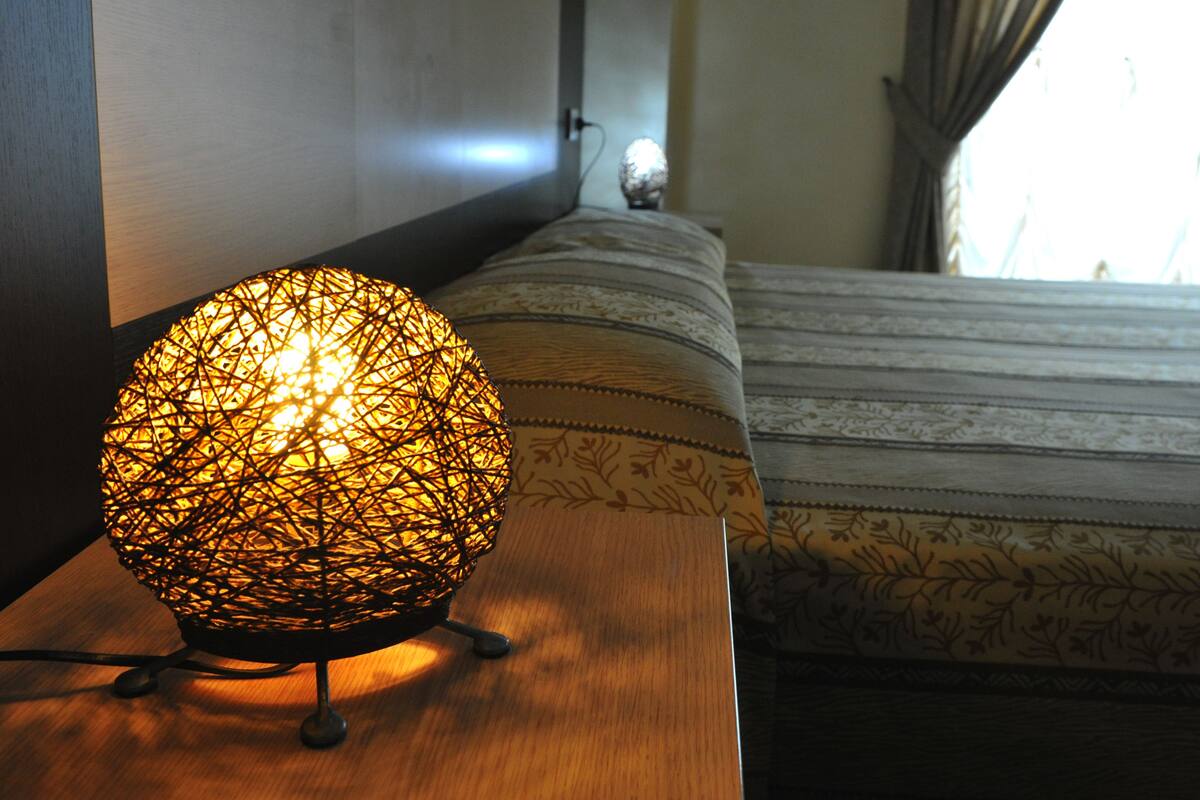 A room with a wooden nightstand, a decorative spherical light, a patterned bedspread, and a view of a window with curtains.