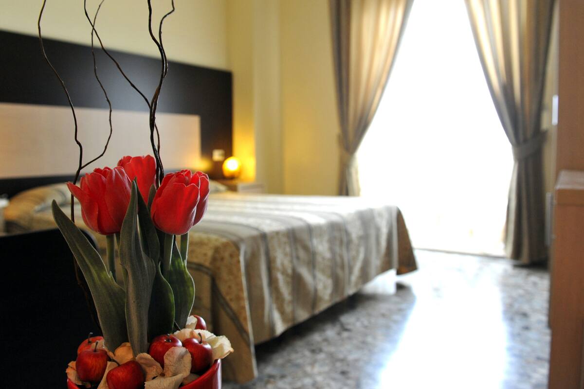 A hotel room with a bed, a vase with red tulips and apples, and a nightstand with a lamp.