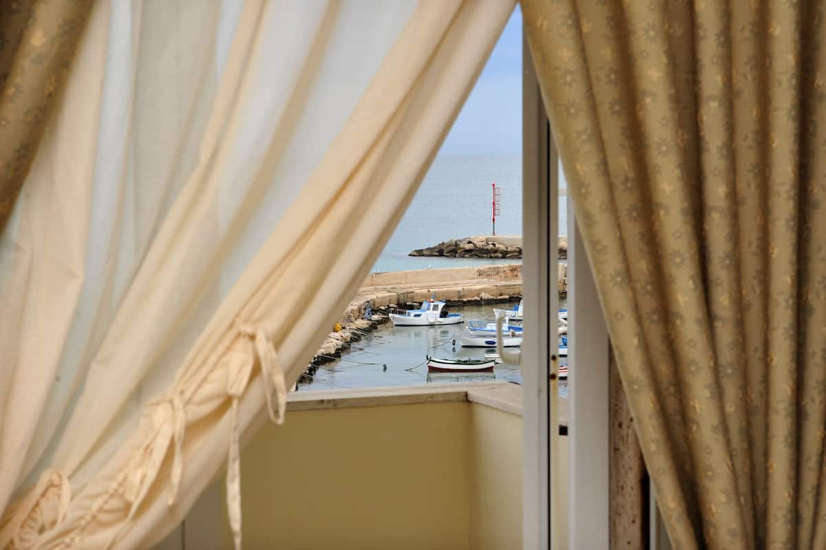 A view through a window with curtains, revealing a marina with boats and a lighthouse in the distance.