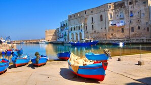 A harbor with several small boats docked, a large historic building with arched windows, and a clear blue sky. - Hotel Gabbiano (Mola di Bari)