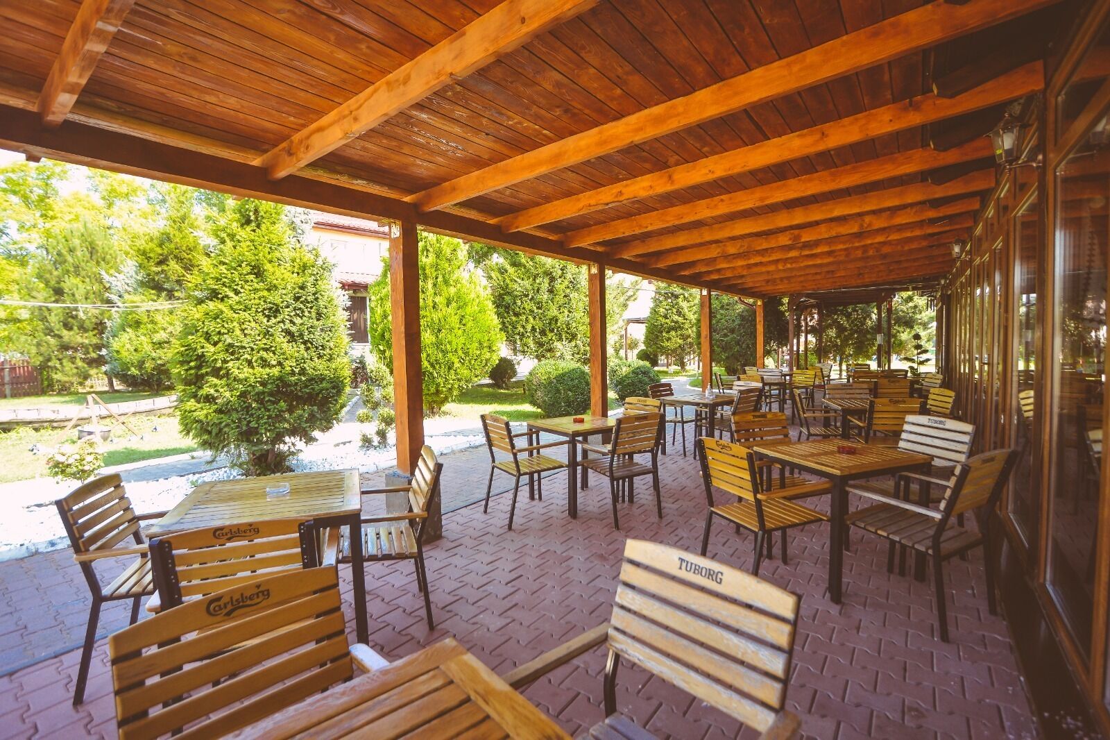 Un espace extérieur couvert, aménagé en patio, avec des tables et des bancs en bois, un sol en briques et une vue sur un jardin et des arbres.