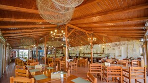 A spacious restaurant with wooden tables and chairs, a high ceiling with hanging lights, and a stone wall in the background.