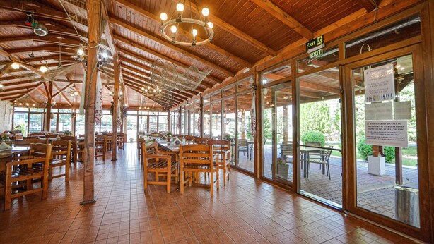 A spacious indoor dining area with wooden tables and chairs, large windows, and a high ceiling with exposed beams.