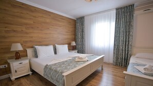 A hotel room with a wooden floor, a bed with white linens, a nightstand with a lamp, and a window with curtains.