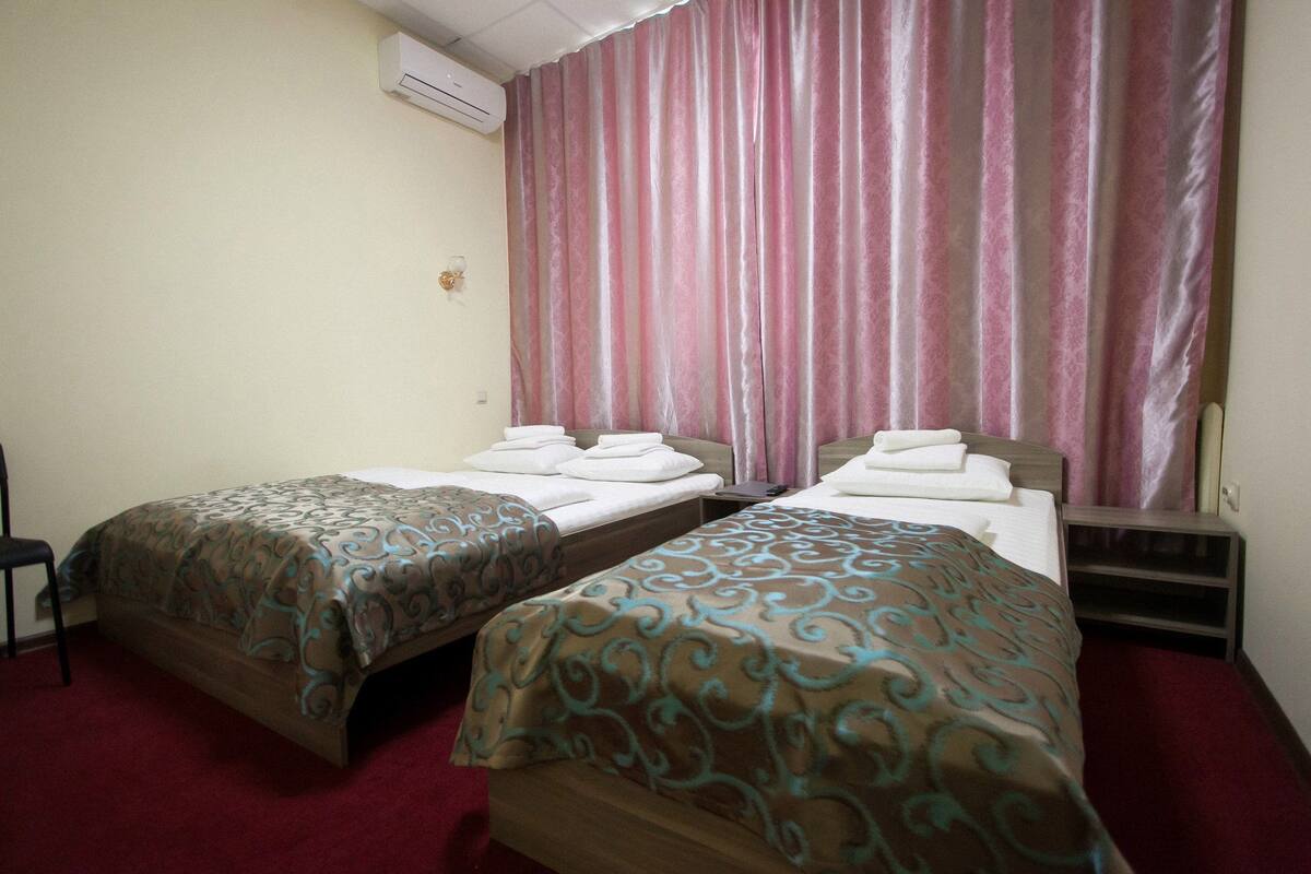 Hotellihuoneessa on kaksi erillistä sänkyä, joissa on kummassakin kuviollinen peitto, yöpöytä ja lamppu. Huoneessa on punainen matto, vaaleanpunaisilla verhoilla varustettu ikkuna ja seinälle asennettu ilmastointilaite.