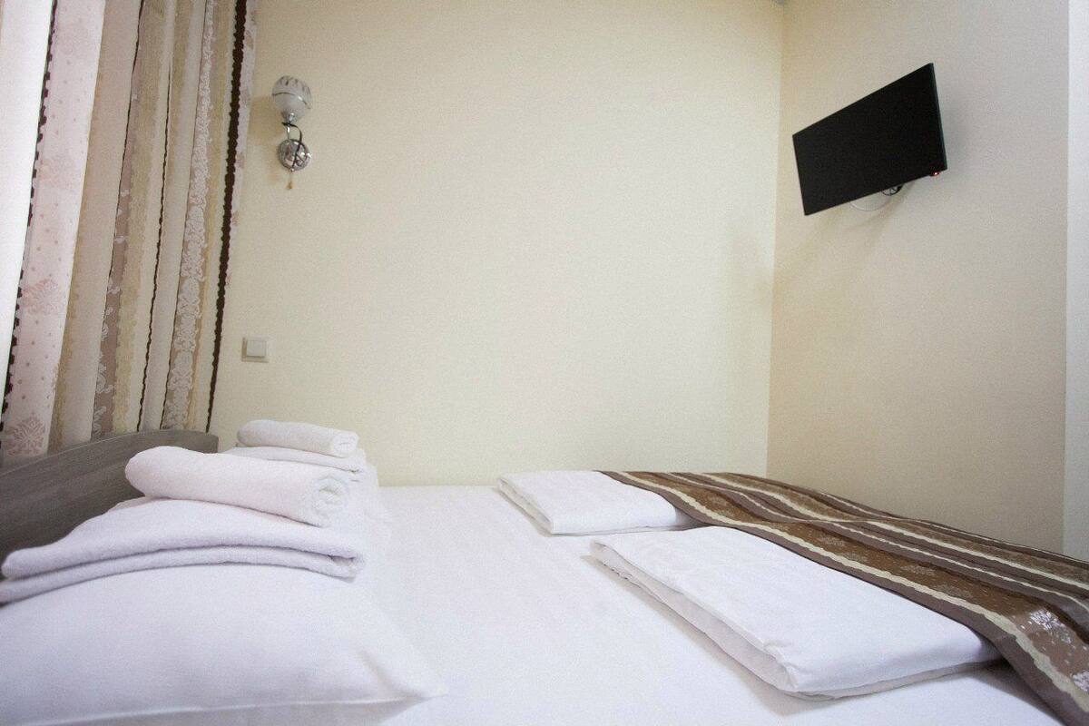 Hotellihuone, jossa on sänky, valkoiset pyyhkeet, seinälle asennettu televisio ja kukkakuviollinen verho.