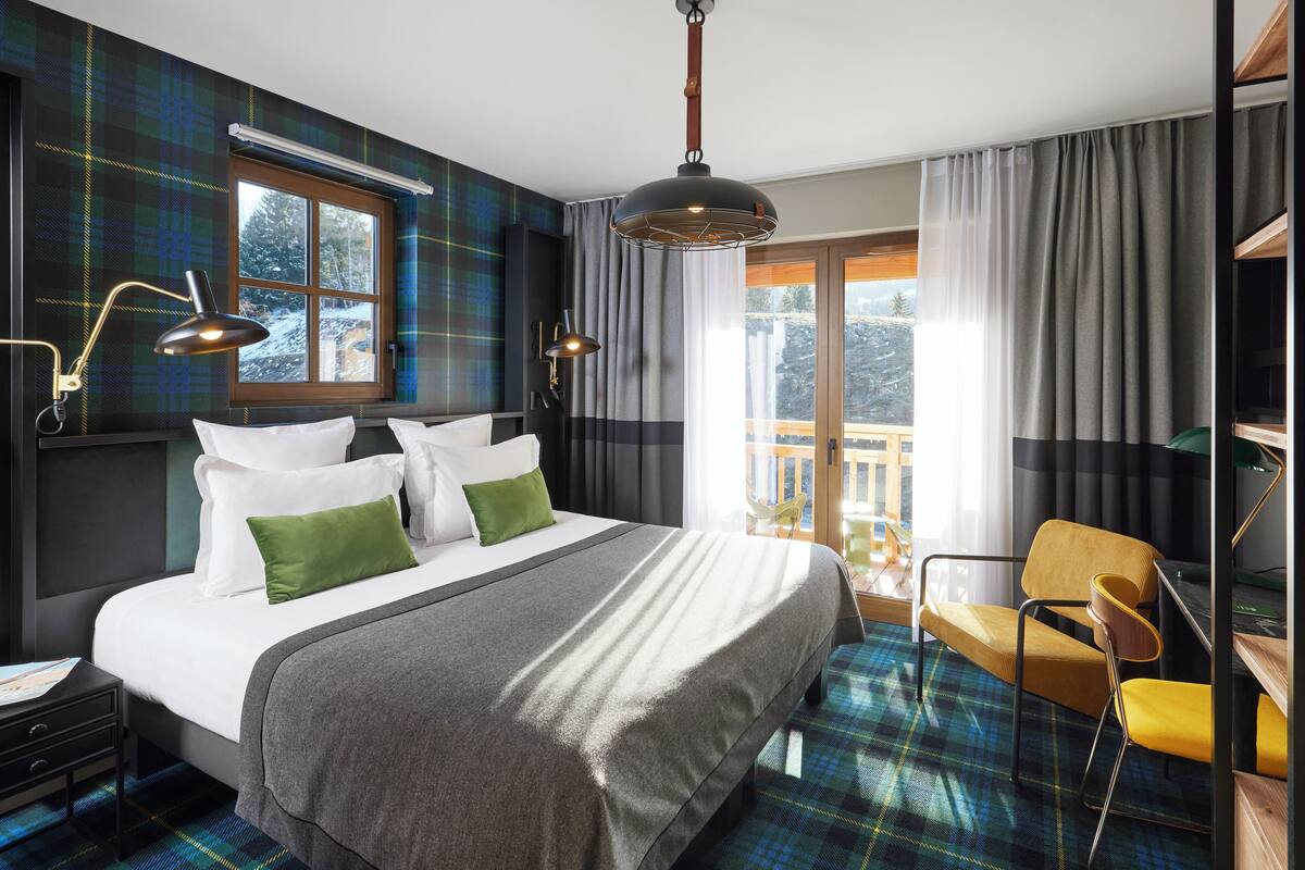 A modern hotel room with a large bed, a desk, a chair, and a balcony with a view of trees.