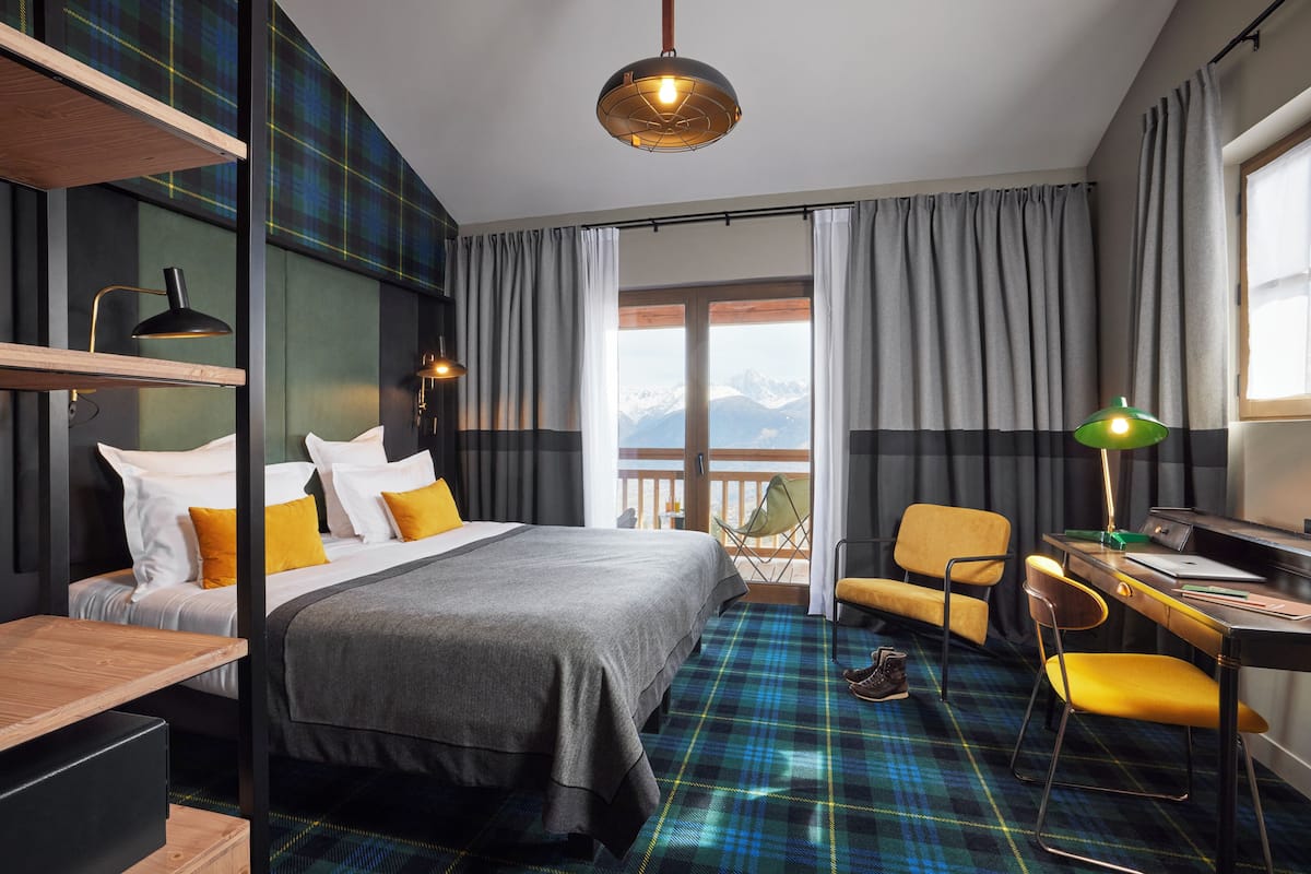 A modern hotel room with a bed, desk, chair, and a view of mountains through the window.