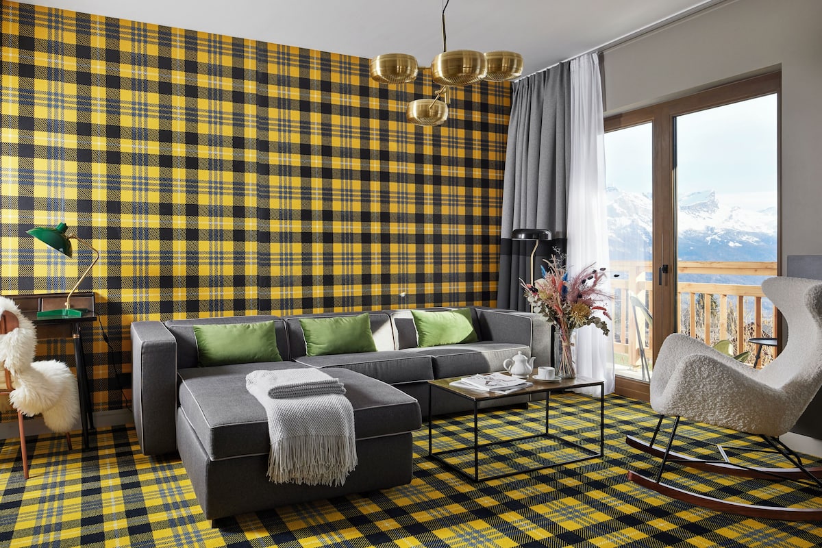 A modern living room with a plaid-patterned wall, a gray sofa with green cushions, a white fluffy chair, and a view of mountains through large windows.