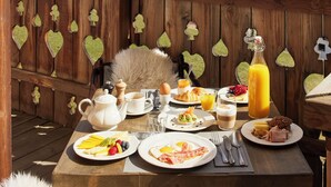 Una terraza de madera con una mesa de desayuno dispuesta para dos, que incluye platos con comida, una cafetera y una botella de zumo de naranja.