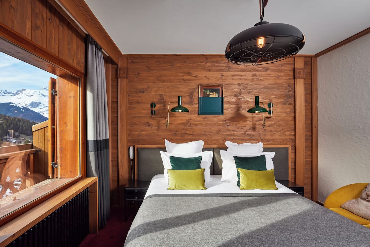 A hotel room with a wooden interior, a bed with white and green pillows, a view of snow-capped mountains, and a black pendant light.