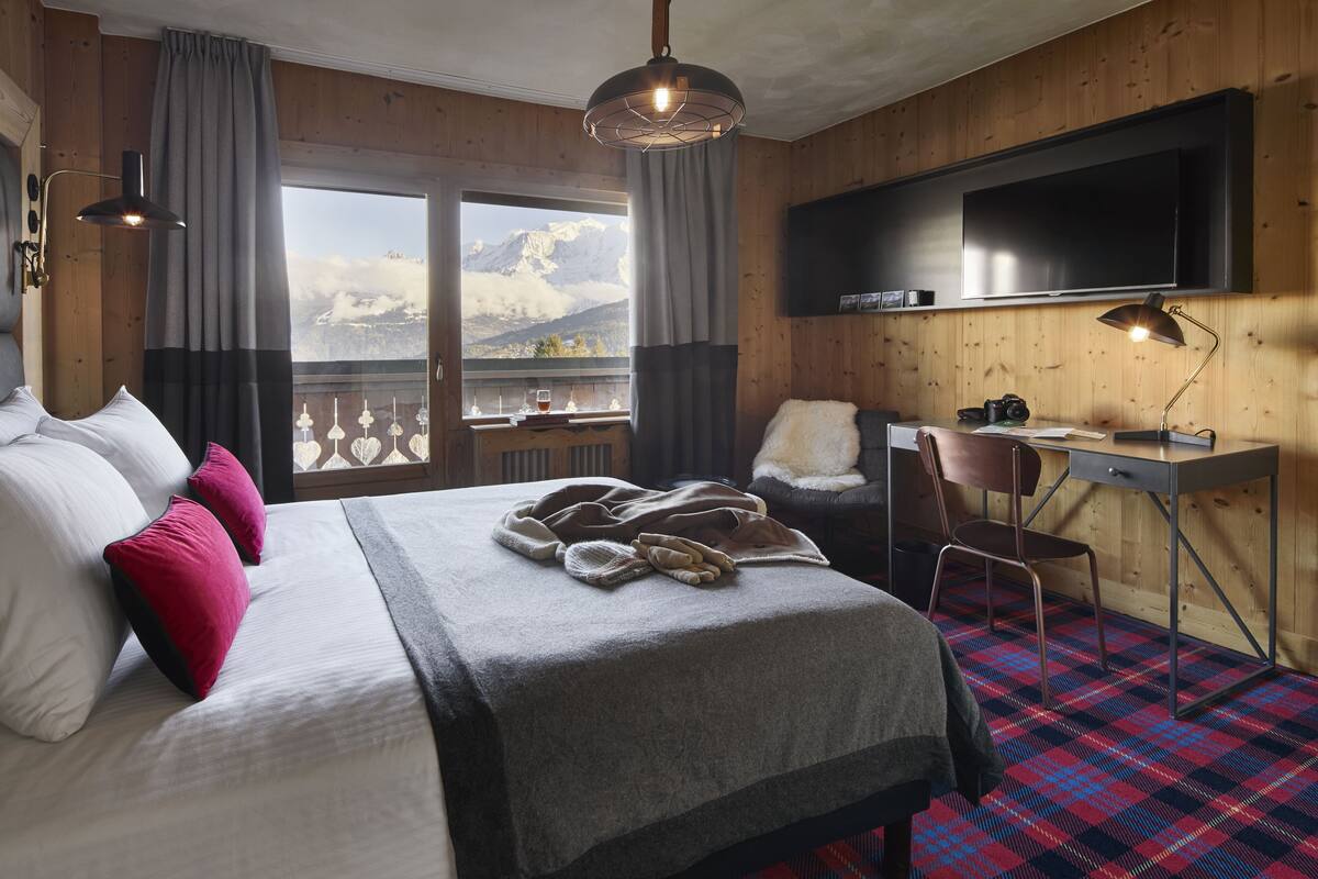 A hotel room with a bed, a desk, a chair, a television, and a view of mountains.