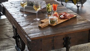 Una mesa de madera rústica con una copa de vino blanco, una copa de vino rosado, una tabla de madera con embutidos y un soporte de metal con botellas y copas.