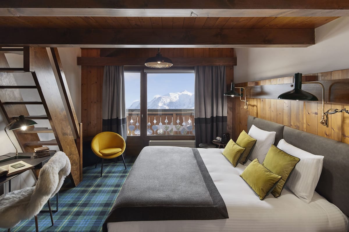 A hotel room with a bed, a desk, a chair, a window with a view of mountains, and wooden walls.