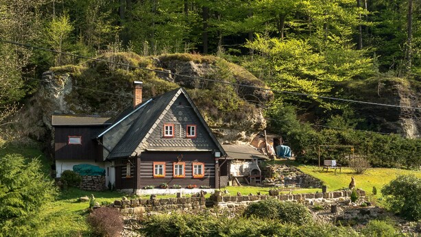 Een traditioneel houten huis met een steil dak, omringd door weelderig groen en een stenen muur.