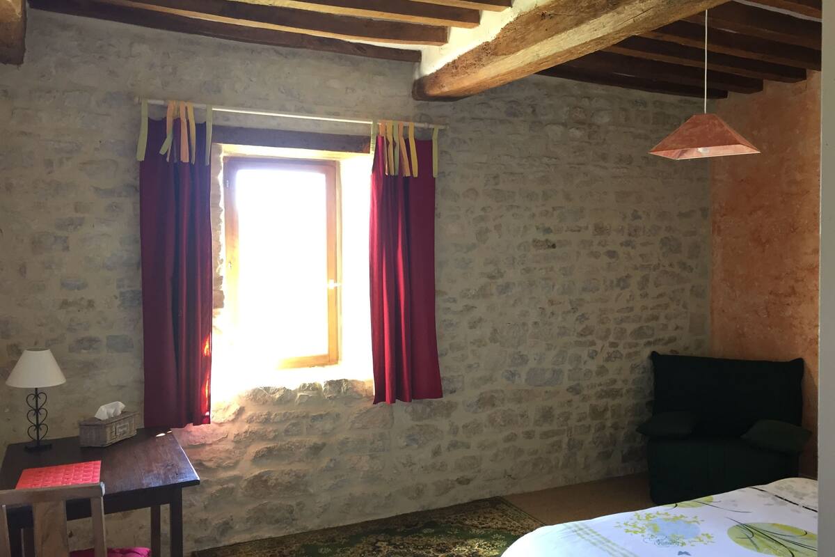 Una habitación rústica con pared de piedra, techo de madera, cama con colcha estampada, sofá verde, mesita con lámpara y ventana con cortinas rojas.