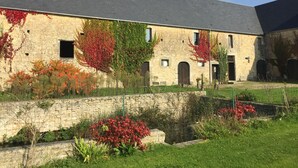 A stone building with a sloped roof, arched doorways, and a courtyard with a stone wall and flowering plants. - La Ferme Manoir Saint Barthélemy (Juaye-Mondaye)