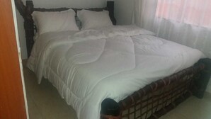 A bed with a wooden frame and white bedding in a room with a plain wall and a window with sheer curtains. - Maurica Homes (Nairobi)