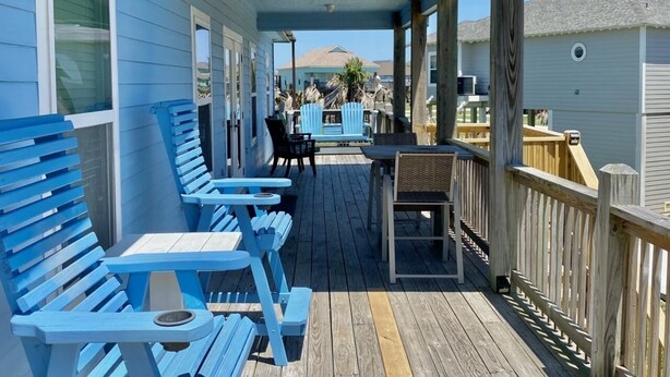 A covered outdoor deck with blue chairs and tables, wooden railings, and a view of the ocean and other buildings. - Paradise Palms - 4 Bedrooms - 2 Baths - Sleeps 10 (Crystal Beach)