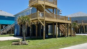A two-story wooden structure with a balcony, supported by stilts, in a residential area with houses and a clear sky. - Paradise Palms - 4 Bedrooms - 2 Baths - Sleeps 10 (Crystal Beach)