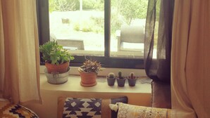 Janela com cortinas, vista para o verde lá fora e uma pequena mesa com plantas e almofadas.