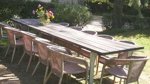 Een buitenterras met een houten tafel en rieten stoelen, omringd door groen en een gebouw met blauwe luiken.