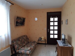 Une pièce avec un canapé à motifs floraux, une petite table avec une lampe, un téléviseur posé sur un meuble en bois et une porte avec une fenêtre.