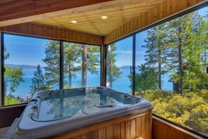 Un bain à remous dans un chalet en bois doté de grandes fenêtres donnant sur un lac et une forêt.