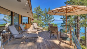 A wooden deck with outdoor seating and a large umbrella, overlooking a lake with trees and mountains in the background. - Anchors Aweigh - Lakefront - TLUXP (Tahoe City)