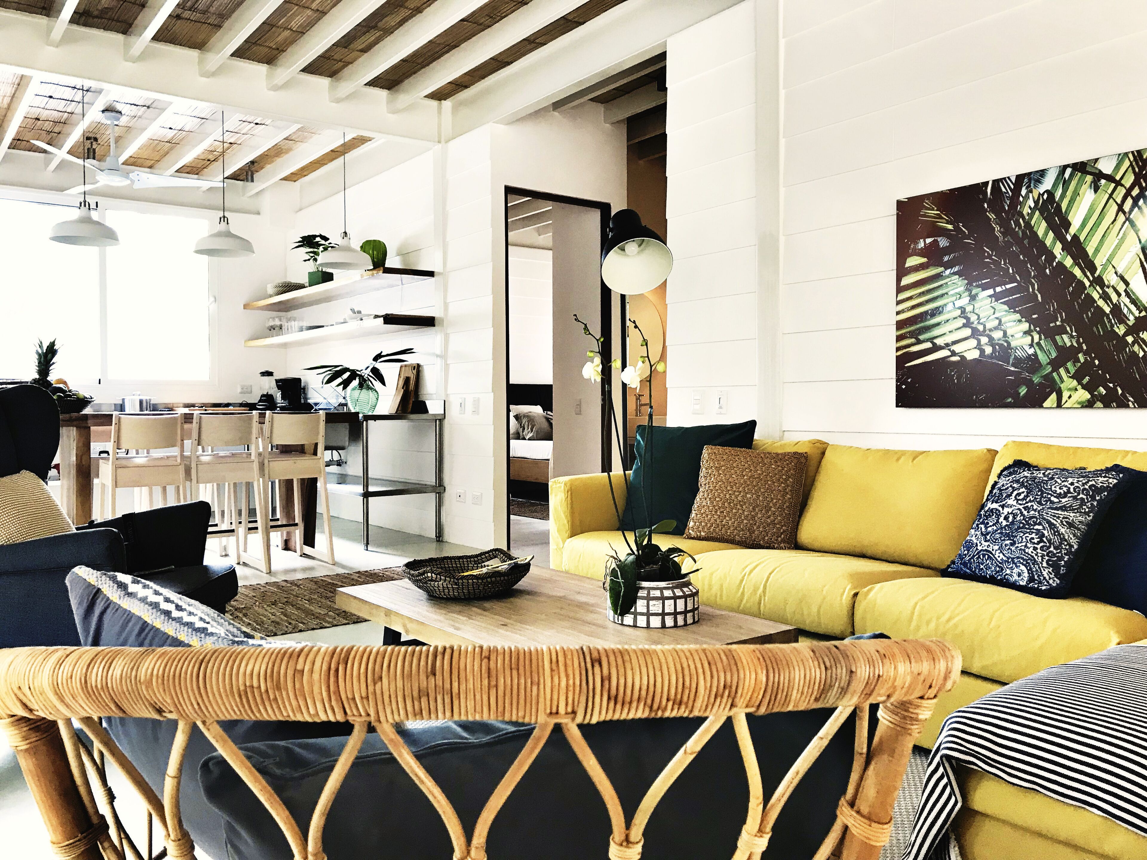 A modern living room with a yellow sofa, wicker chairs, and a dining area with a wooden table and chairs.