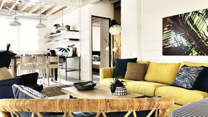 Een moderne woonkamer met een gele bank, rieten stoelen en een eethoek met een houten tafel en stoelen.