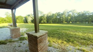 Un espacio al aire libre cubierto con pilares de ladrillo, suelo de grava y un campo con árboles de fondo.
