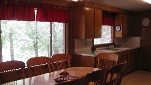 A wooden dining room with a table and chairs, a kitchen with wooden cabinets, a window with red curtains, and a clock on the wall.