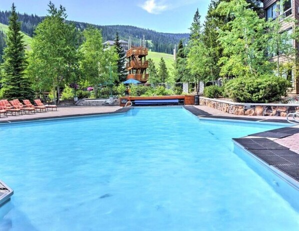 Piscina con sillones de descanso, rodeada de árboles y montañas al fondo.