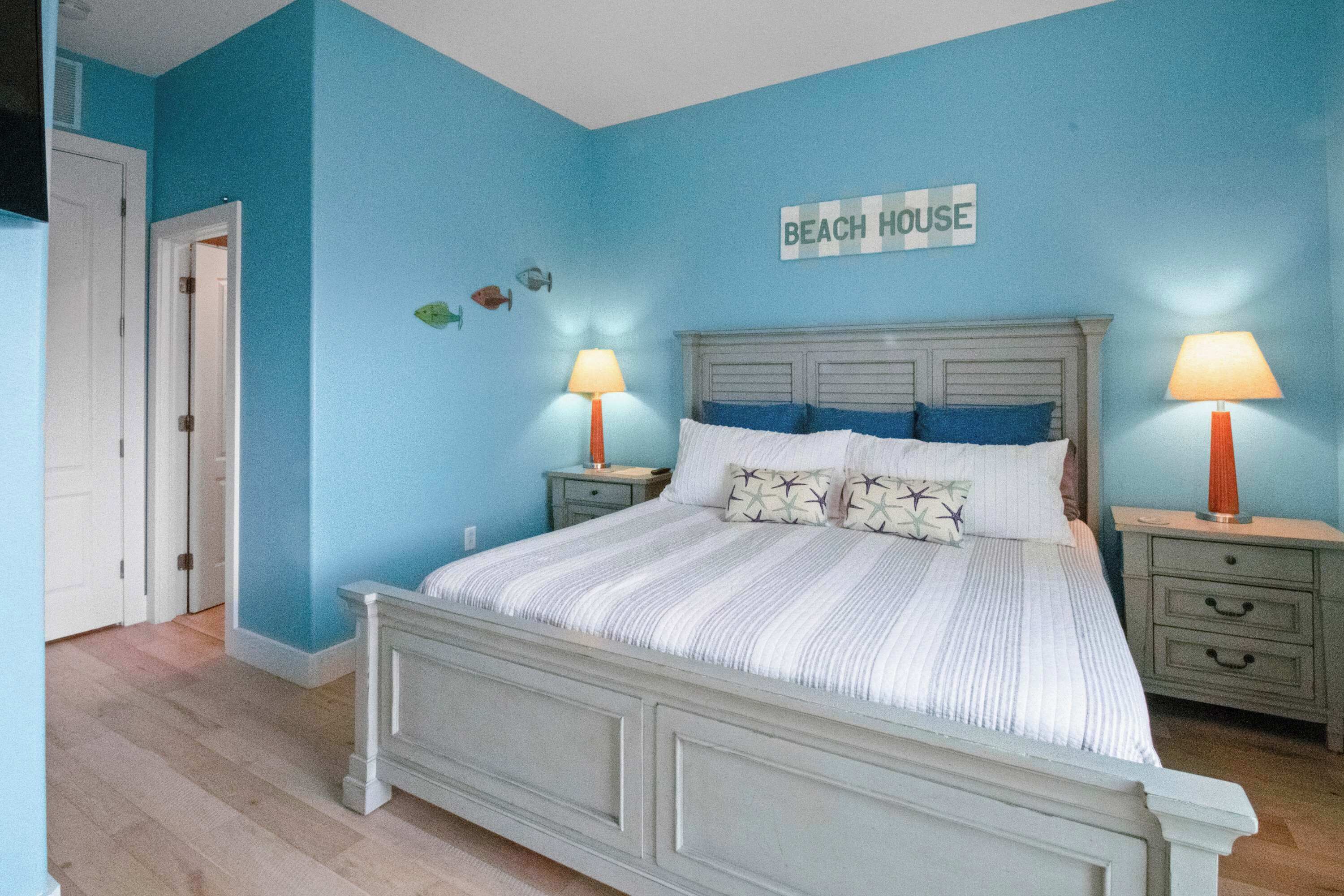 A bedroom with a bed, two bedside tables, and a wall sign reading "BEACH HOUSE".