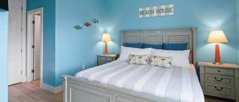 A bedroom with a bed, two bedside tables, and a wall sign reading "BEACH HOUSE".