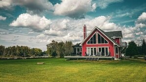 Een groot, rood, twee verdiepingen tellend huis met een opvallende schoorsteen, omgeven door een verzorgd gazon en bomen.
