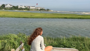 Une personne est assise à une table de pique-nique en bois, près d’une étendue d’eau, avec une vue sur la côte et des bâtiments en arrière-plan.